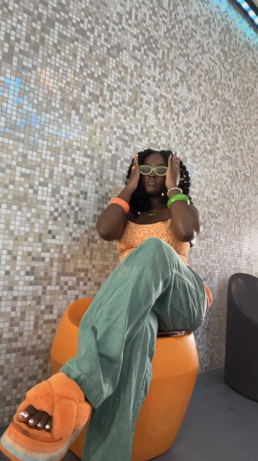 Nelle in an orange floral top, green trousers and fuzzy orange slides against a mosaic wall